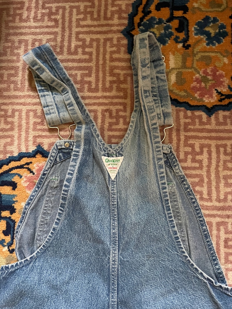 May include: A pair of light blue denim dungarees with adjustable straps. The dungarees have a brand label that reads "OshKosh B'Gosh" and "Vest-Tex".