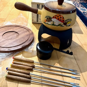 May include: A vintage fondue set with a yellow pot featuring mushroom illustrations, a black stand, wooden-handled forks with colored tips, a wooden serving tray, and a black burner. The set includes a lid and a ladle.