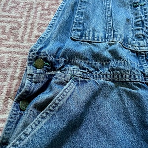 May include: A close-up of a pair of blue denim dungarees with multiple pockets and metal buttons. The dungarees are made of a soft, worn denim fabric.