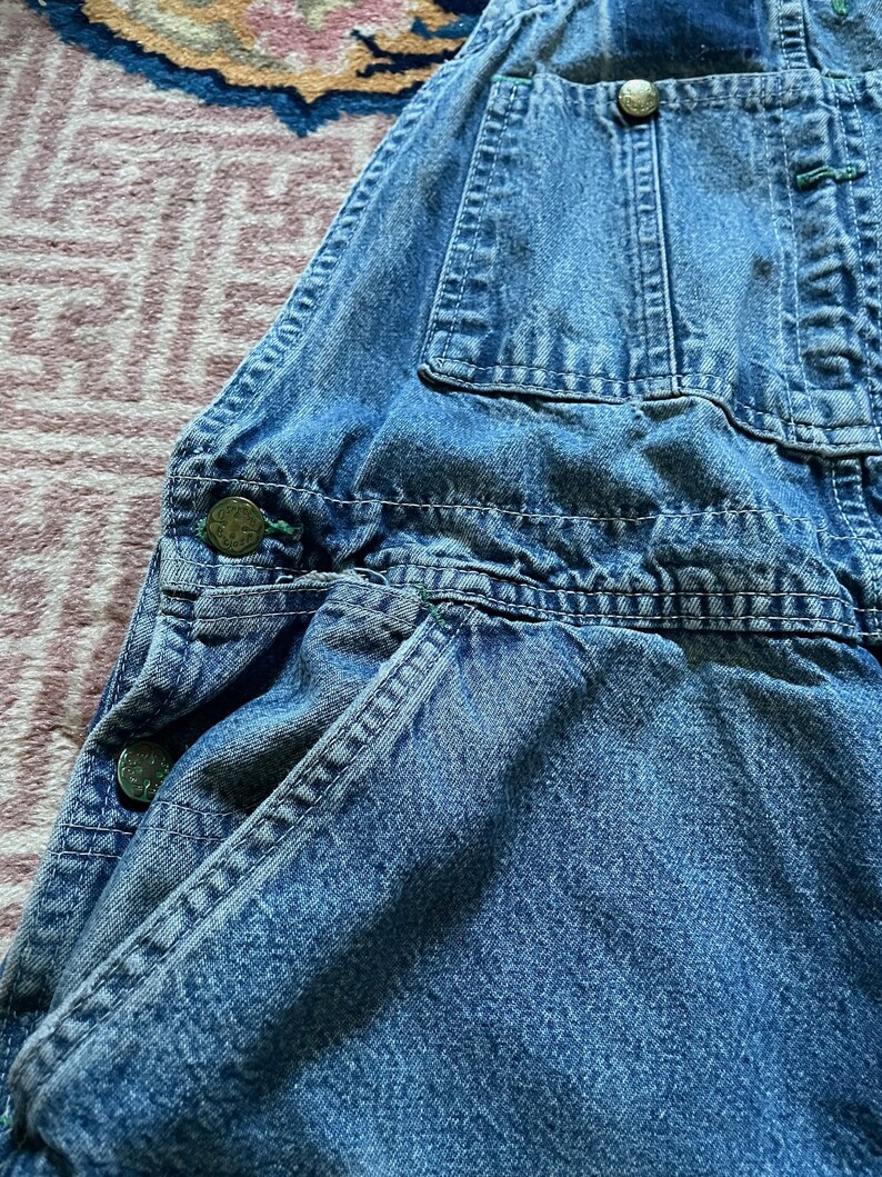 May include: A close-up of a pair of blue denim dungarees with multiple pockets and metal buttons. The dungarees are made of a soft, worn denim fabric.
