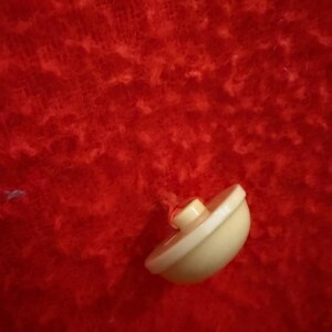 May include: A small, off-white button with a rounded shape and a raised center. The button is set against a vibrant red, textured background, creating a striking contrast. The button is likely for clothing or crafts.