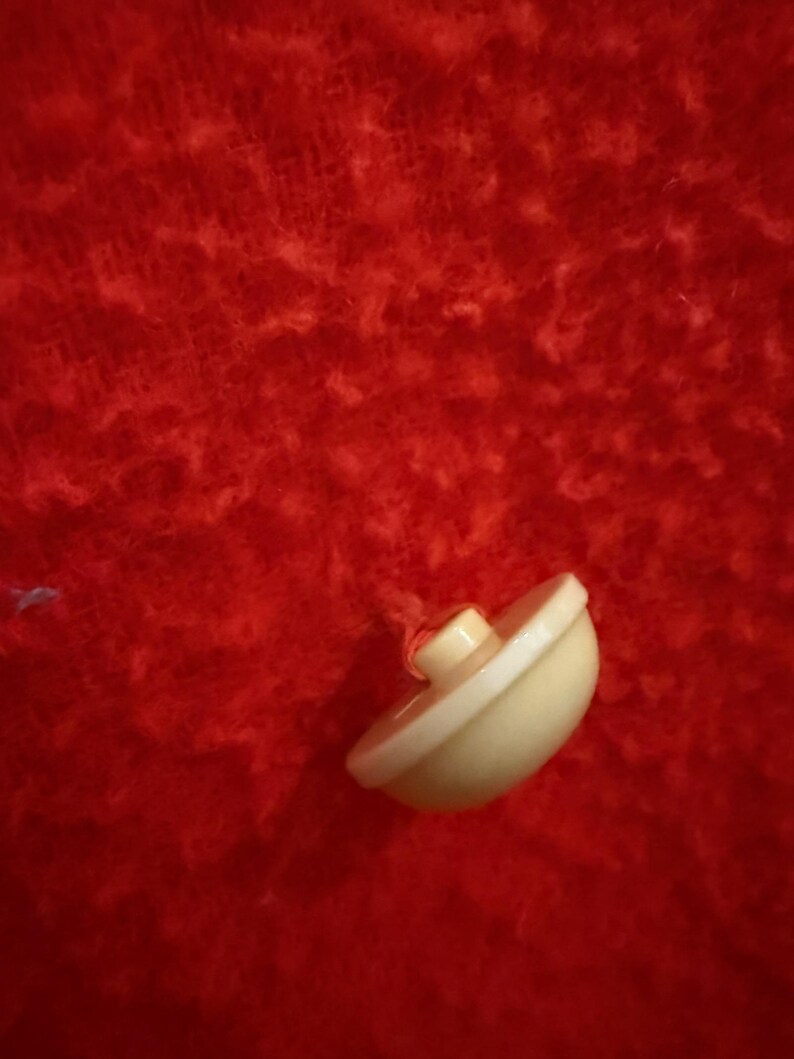 May include: A small, off-white button with a rounded shape and a raised center. The button is set against a vibrant red, textured background, creating a striking contrast. The button is likely for clothing or crafts.