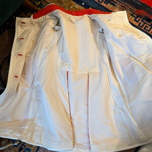 May include: Open view of a white coat with a red collar and red stitching. The coat has red buttons and a white interior. The coat appears to be made of a smooth, possibly water-resistant material. The coat is open, revealing the interior lining.