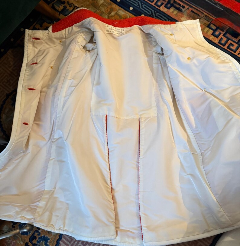 May include: Open view of a white coat with a red collar and red stitching. The coat has red buttons and a white interior. The coat appears to be made of a smooth, possibly water-resistant material. The coat is open, revealing the interior lining.