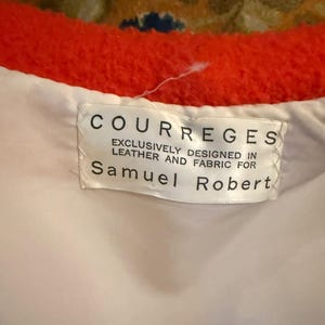 May include: Close-up of a label from a Courreges garment. The label is white with black text that reads "COURREGES EXCLUSIVELY DESIGNED IN LEATHER AND FABRIC FOR Samuel Robert". The garment has a bright orange trim.