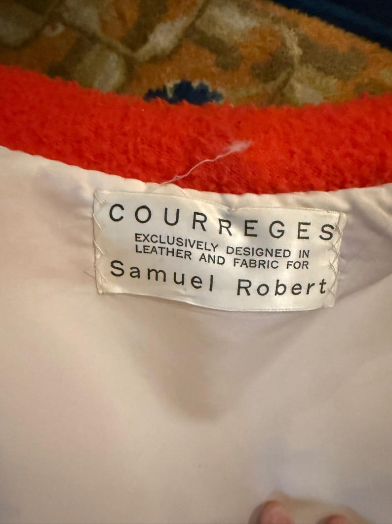 May include: Close-up of a label from a Courreges garment. The label is white with black text that reads "COURREGES EXCLUSIVELY DESIGNED IN LEATHER AND FABRIC FOR Samuel Robert". The garment has a bright orange trim.