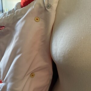 May include: Close-up of a white jacket interior with a bright orange, fuzzy collar. The jacket has a light pink lining and is fastened with two yellow buttons and a silver snap. The jacket is displayed on a mannequin.