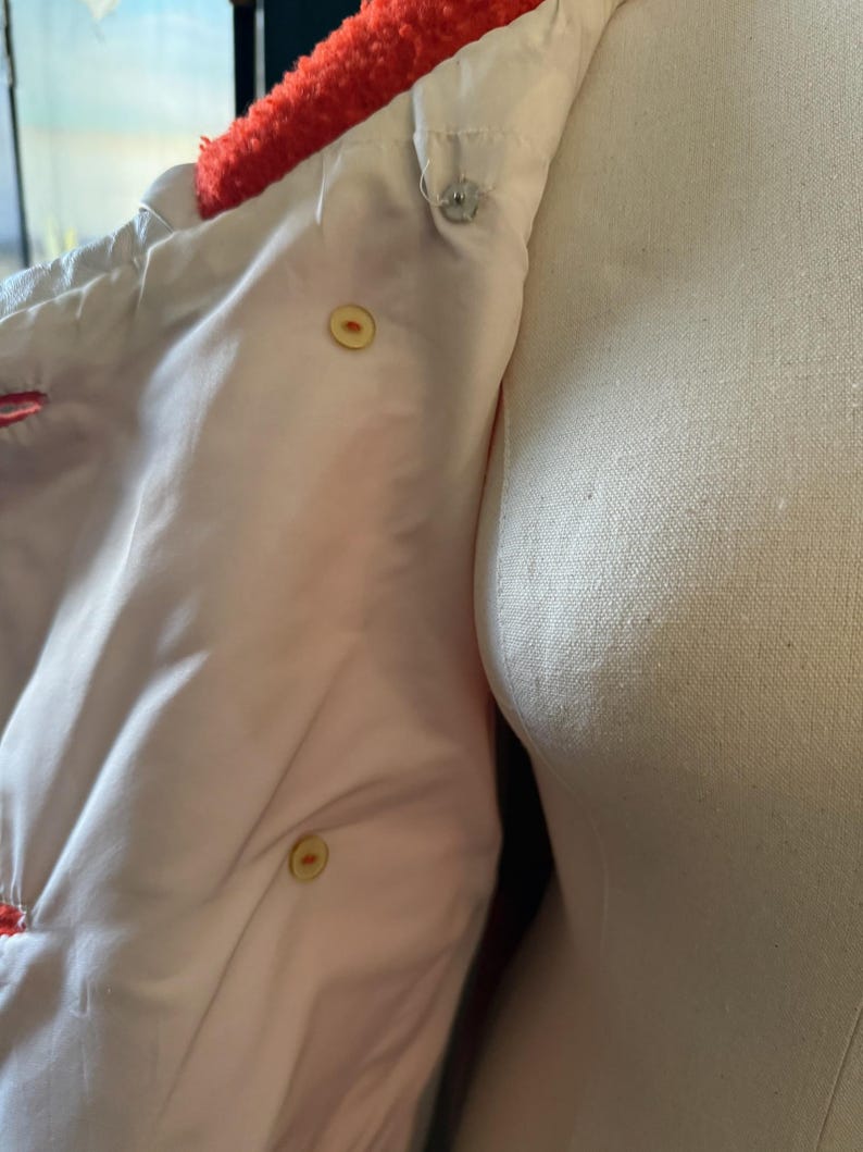 May include: Close-up of a white jacket interior with a bright orange, fuzzy collar. The jacket has a light pink lining and is fastened with two yellow buttons and a silver snap. The jacket is displayed on a mannequin.