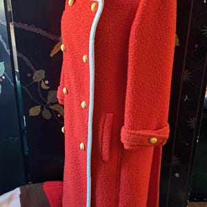 May include: A vibrant red vintage coat with a textured, boucl&eacute; fabric. The coat features a white trim along the collar and edges, and is accented with gold-colored buttons. The long coat has a double-breasted design and cuffed sleeves.
