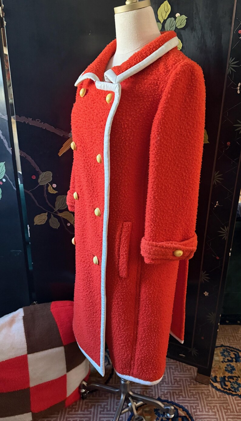 May include: A vibrant red vintage coat with a textured, boucl&eacute; fabric. The coat features a white trim along the collar and edges, and is accented with gold-colored buttons. The long coat has a double-breasted design and cuffed sleeves.