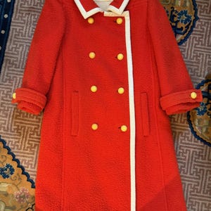 May include: A vintage red coat with a white trim and yellow buttons. The coat has a double-breasted design, a white collar, and cuffed sleeves. The label reads "COURREGES".