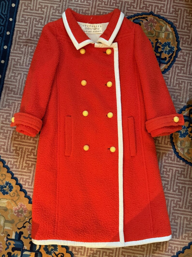 May include: A vintage red coat with a white trim and yellow buttons. The coat has a double-breasted design, a white collar, and cuffed sleeves. The label reads "COURREGES".