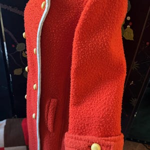 May include: A vibrant red coat with a textured, boucl&eacute; fabric. The coat features a white trim along the collar and front, accented by yellow buttons. The sleeve cuffs are also detailed with yellow buttons, offering a classic, tailored look.