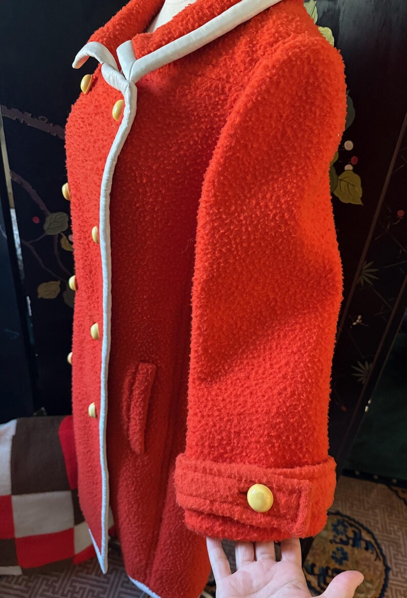 May include: A vibrant red coat with a textured, boucl&eacute; fabric. The coat features a white trim along the collar and front, accented by yellow buttons. The sleeve cuffs are also detailed with yellow buttons, offering a classic, tailored look.