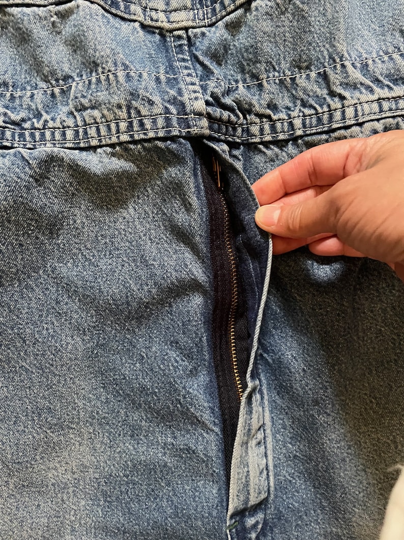 May include: Close-up of a blue denim zipper with a gold zipper pull. The zipper is partially open and the fabric is slightly creased.