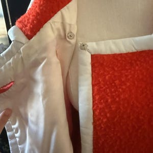 May include: Close-up of a vintage coat with a vibrant orange textured exterior and white trim. The coat features a satin-like white lining and decorative silver buttons. The collar and cuffs are trimmed with the orange textured material.