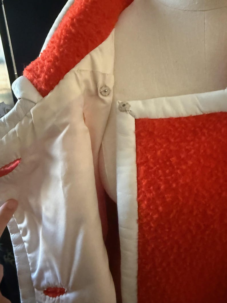 May include: Close-up of a vintage coat with a vibrant orange textured exterior and white trim. The coat features a satin-like white lining and decorative silver buttons. The collar and cuffs are trimmed with the orange textured material.