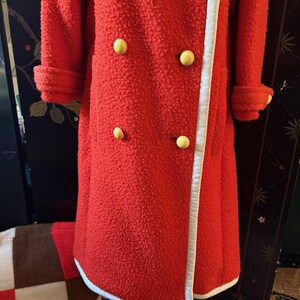 May include: A vintage red coat with a textured surface, featuring large yellow buttons and white trim along the edges. The coat has long sleeves with folded cuffs. A retro fashion item.