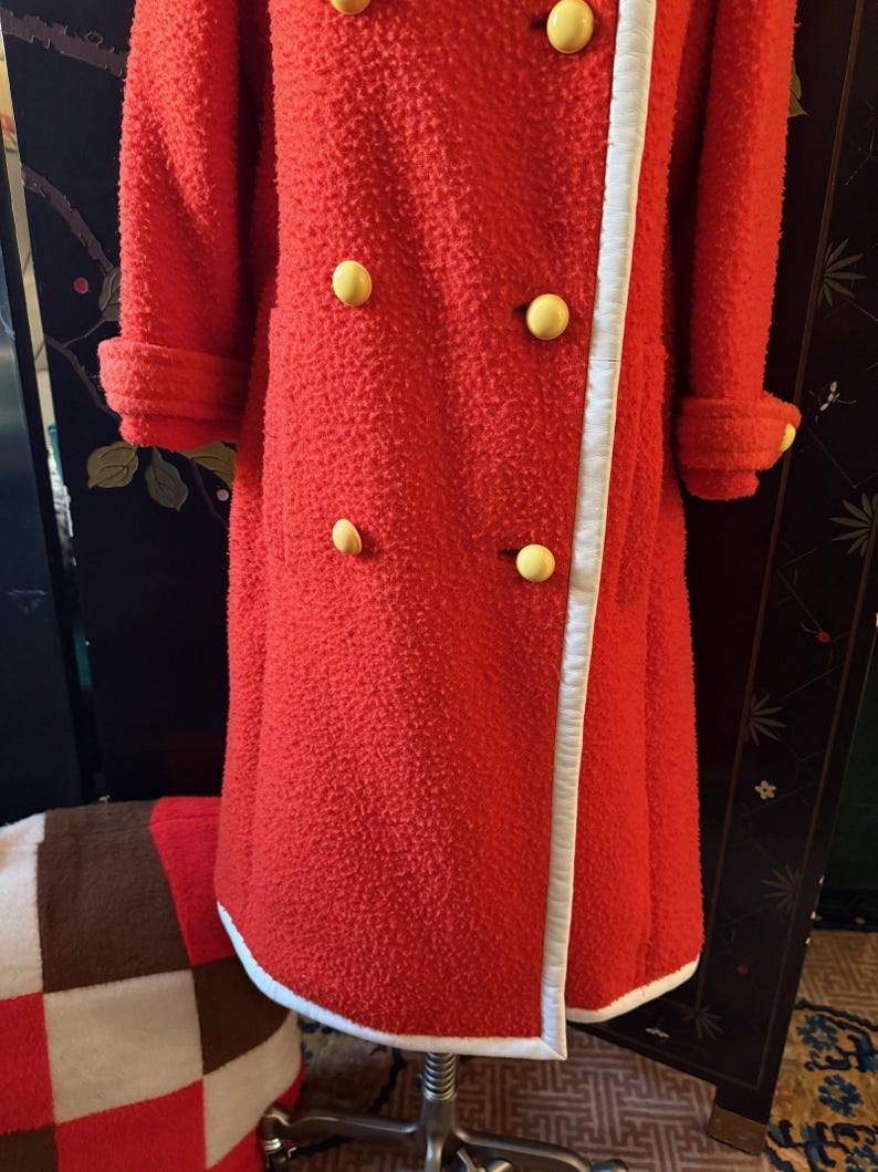 May include: A vintage red coat with a textured surface, featuring large yellow buttons and white trim along the edges. The coat has long sleeves with folded cuffs. A retro fashion item.