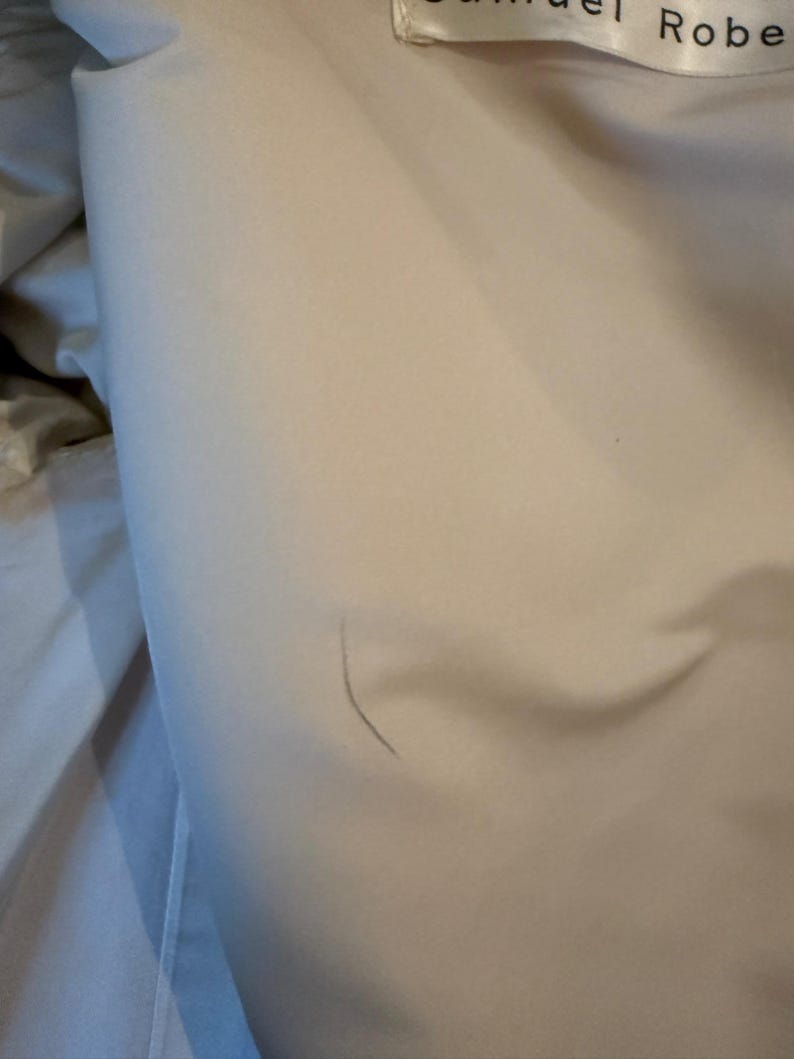 May include: Close-up of a light beige fabric with a subtle sheen. The fabric is slightly wrinkled, with a visible seam and the text "Samuel Robe" on a label.