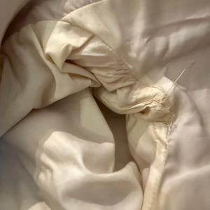 May include: Close-up of a cream-colored fabric, possibly a garment lining, with visible stitching and loose threads. The fabric appears soft and slightly wrinkled, with subtle variations in tone.