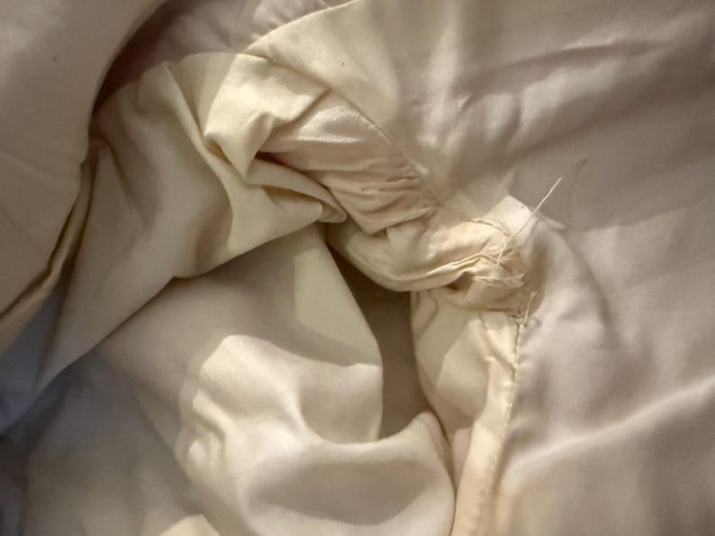 May include: Close-up of a cream-colored fabric, possibly a garment lining, with visible stitching and loose threads. The fabric appears soft and slightly wrinkled, with subtle variations in tone.