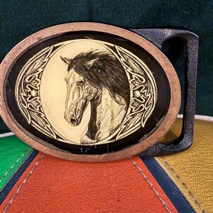 May include: Oval-shaped belt buckle with a detailed horse head design. The buckle features a copper-colored border, a black center, and a gold-toned horse illustration. The buckle's hardware is black.