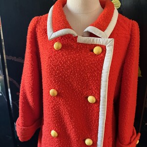 May include: A bright red vintage coat with a textured fabric. The coat features a white trim along the collar and front edges, and is accented with large yellow buttons. The sleeves are cuffed, and the coat is displayed on a mannequin.