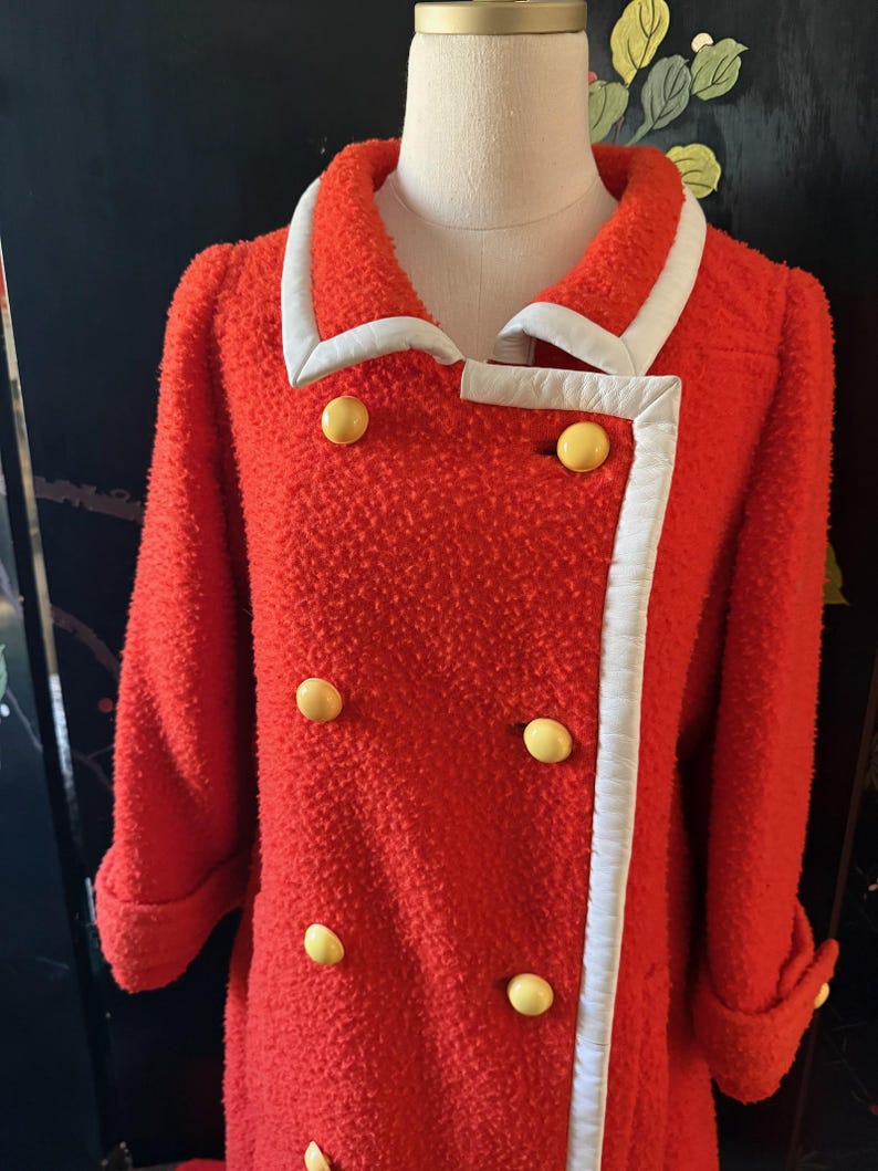 May include: A bright red vintage coat with a textured fabric. The coat features a white trim along the collar and front edges, and is accented with large yellow buttons. The sleeves are cuffed, and the coat is displayed on a mannequin.