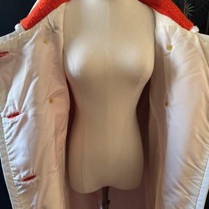 May include: A white coat with a bright orange, textured collar and red stitching details. The coat is buttoned with gold-colored buttons and is displayed on a mannequin. The coat's interior is a light pink color.
