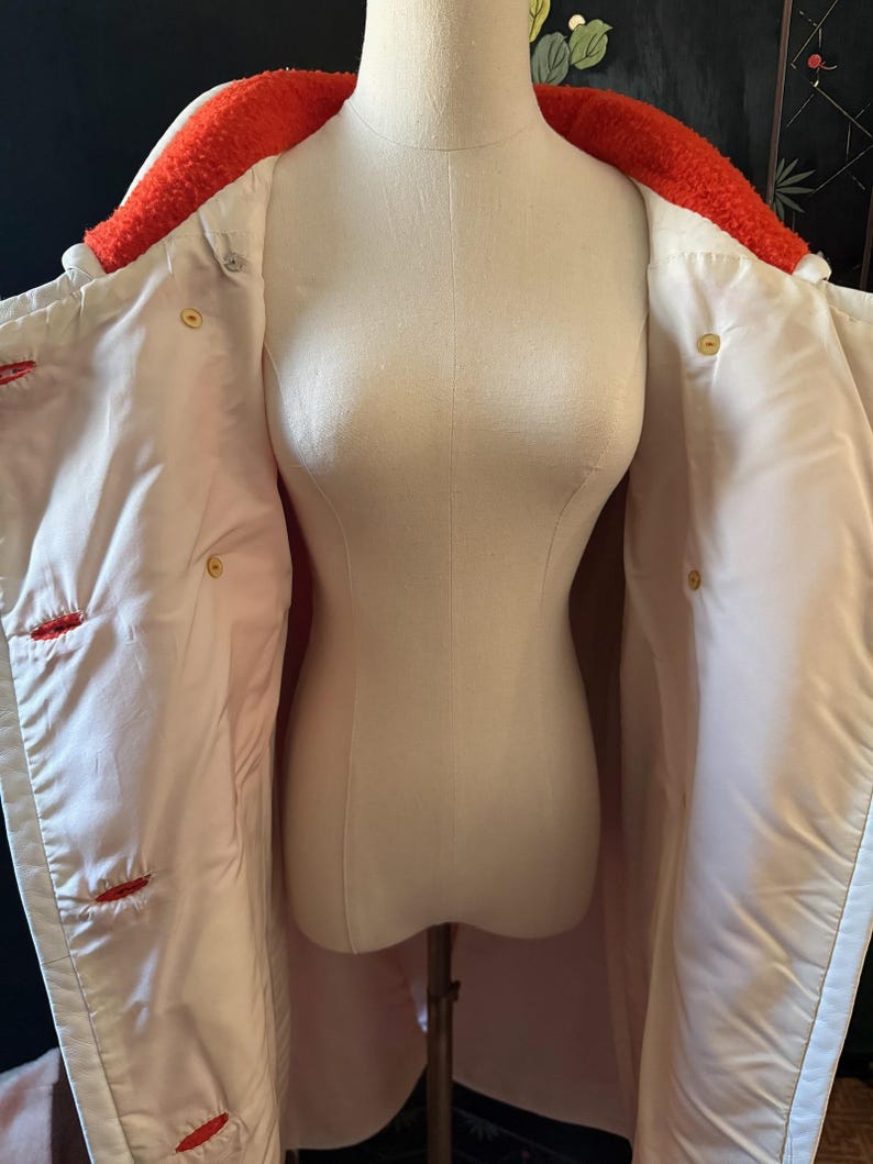 May include: A white coat with a bright orange, textured collar and red stitching details. The coat is buttoned with gold-colored buttons and is displayed on a mannequin. The coat's interior is a light pink color.