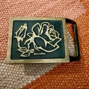 May include: A rectangular belt buckle with a brass frame and a dark teal center. The center features a gold-colored rose design with a bud and leaves. The buckle has a brass loop for the belt.
