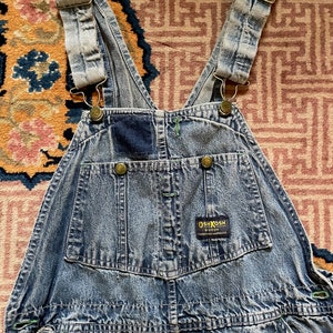 May include: Light blue denim dungarees with adjustable straps and a patch on the front that says "OSHKOSH B'Gosh" in white text.