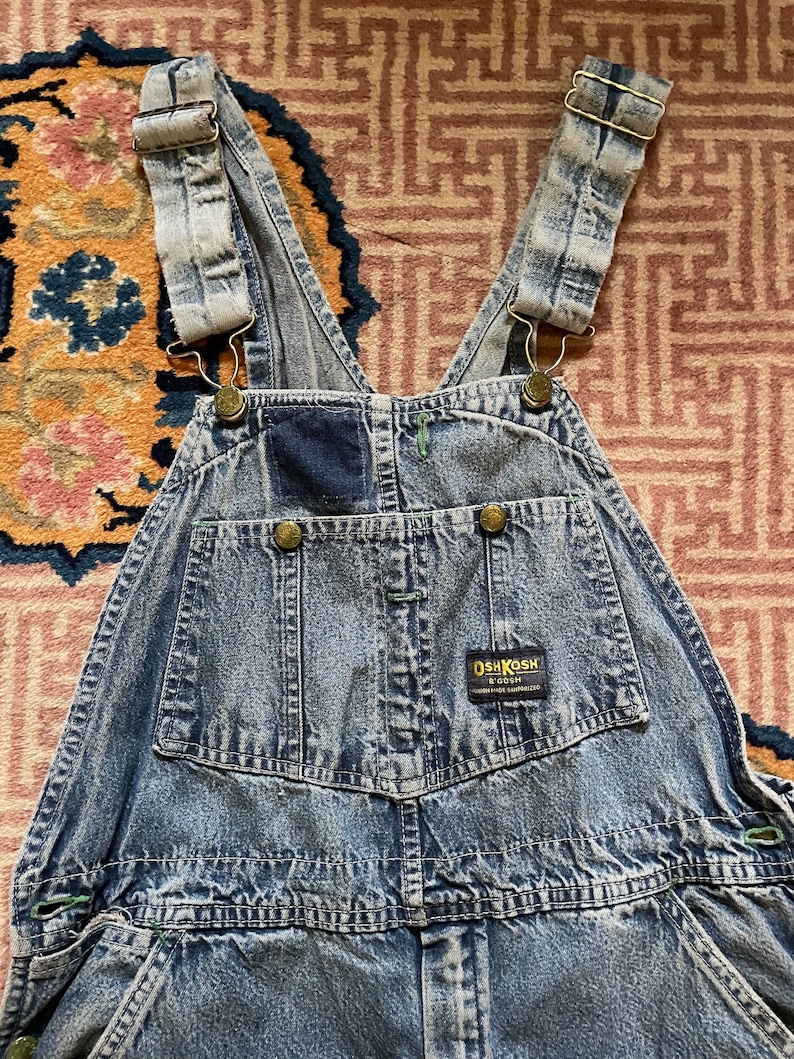 May include: Light blue denim dungarees with adjustable straps and a patch on the front that says "OSHKOSH B'Gosh" in white text.
