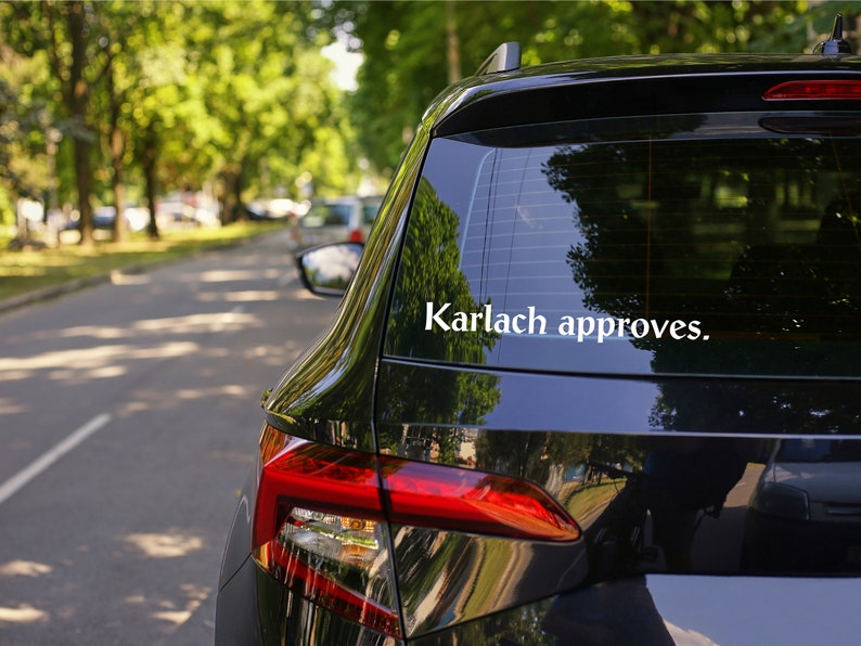 May include: A black car with a white sticker on the back window that says "Karlach approves."