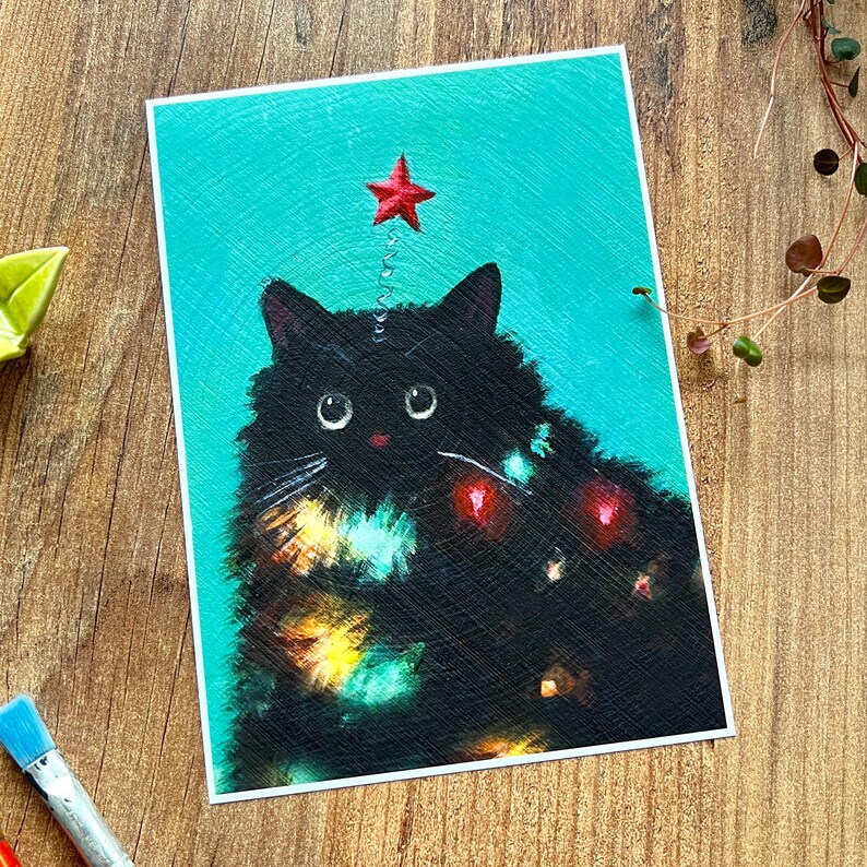 Puede incluir: Un gato negro con una estrella roja en la cabeza est&aacute; decorado con luces de colores, que se asemejan a un &aacute;rbol de Navidad. El gato est&aacute; sentado sobre un fondo turquesa.