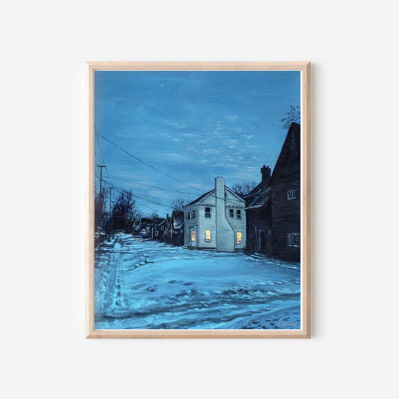 May include: A framed print depicting a snow-covered street at night. A white house with illuminated windows is the focal point. The artwork is set against a dark blue sky and framed with a light wood border.