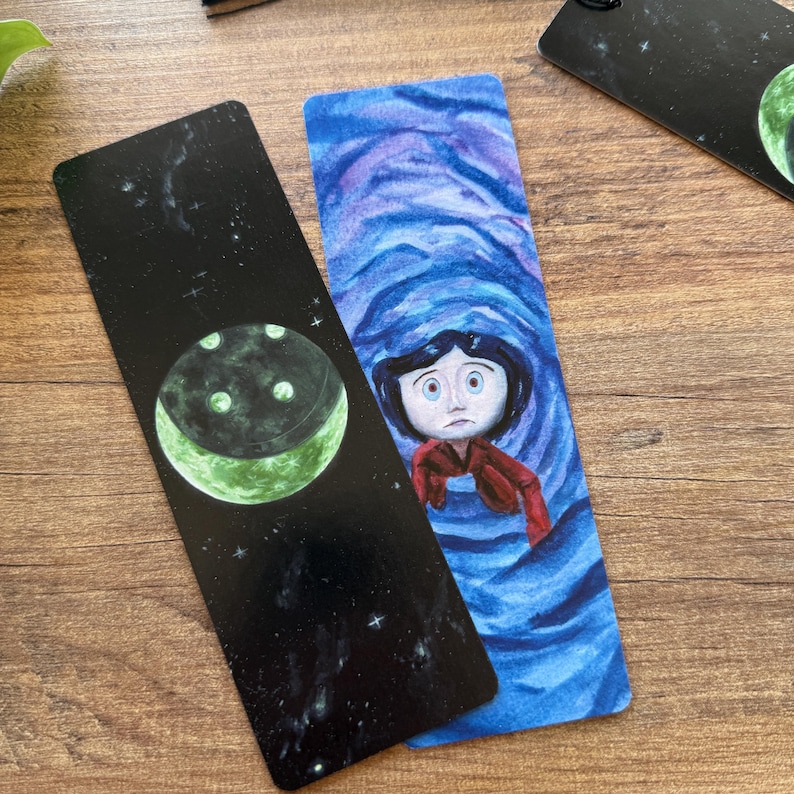以下が含まれることがあります： 2つのしおり。1つは、星が散りばめられた黒い背景に緑色の月が描かれたもの。もう1つは、青い渦巻く水の中に赤い髪の女の子が描かれた水彩画です。