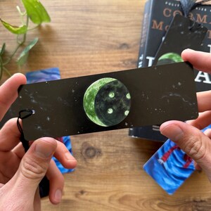 以下が含まれることがあります： 黒い背景に白い星が散りばめられた、緑色の三日月と白いクレーターが描かれた黒いしおり。