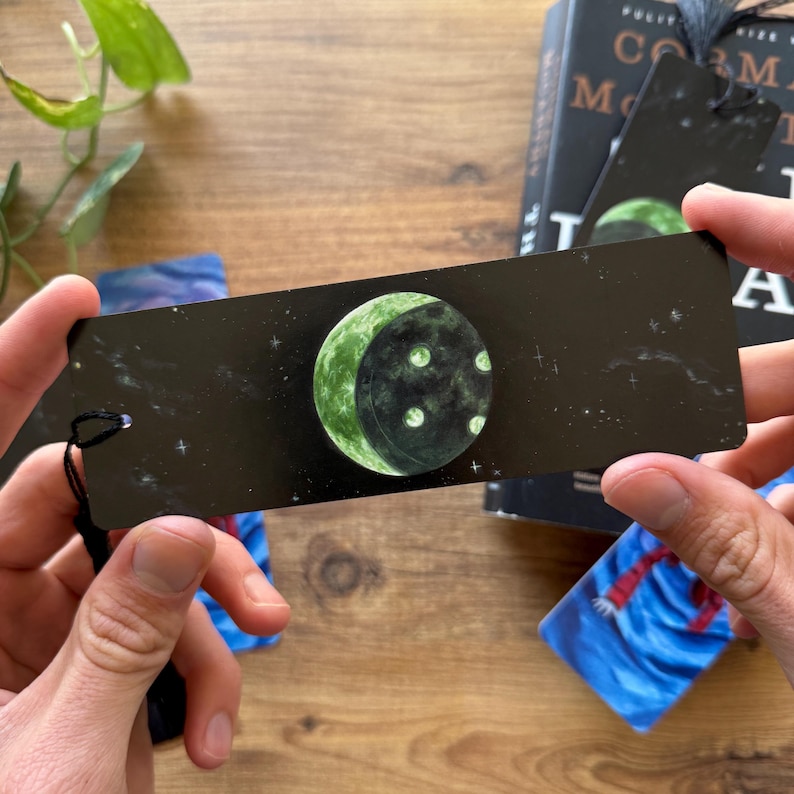 以下が含まれることがあります： 黒い背景に白い星が散りばめられた、緑色の三日月と白いクレーターが描かれた黒いしおり。