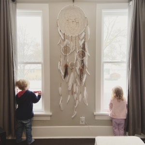 May include: A large, cream-colored dreamcatcher with multiple smaller dreamcatchers and white feathers hanging below. The dreamcatcher is hanging on a light gray wall between two windows. Two children are looking out the windows.