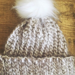 Puede incluir: Un gorro tejido a mano en un color neutro con un pompón blanco esponjoso en la parte superior. El gorro está hecho de hilo grueso, con un borde acanalado y un patrón de tejido texturizado. Un gorro de invierno cálido.