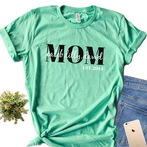 Pu&ograve; includere: Una maglietta verde chiaro con la parola "MOM" in lettere nere e il testo "caleb.lilly.david EST. 2014" in lettere bianche.