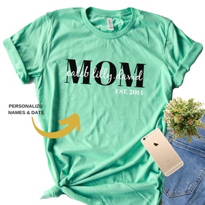 Pu&ograve; includere: Una maglietta turchese con la parola "MOM" in lettere maiuscole nere e i nomi "Caleb, Lilly, David" scritti in corsivo bianco sotto. Il testo "EST. 2014" &egrave; scritto in nero sotto i nomi.