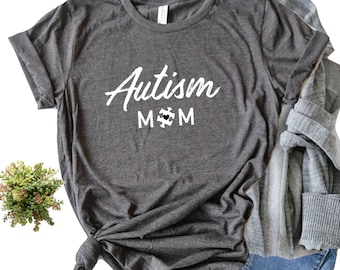 May include: Gray t-shirt with the text "Autism Mom" in white lettering. A white puzzle piece with a black heart is in the middle of the word "Mom".