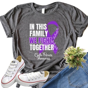 Puede incluir: Una camiseta gris con el texto "In this family we fight together" y una cinta morada con pequeños corazones. El texto "Cystic Fibrosis Awareness" también está en la camiseta.