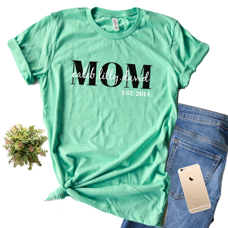 Pu&ograve; includere: Una maglietta verde chiaro con la parola "MOM" in lettere nere e il testo "caleb.lilly.david EST. 2014" in lettere bianche.