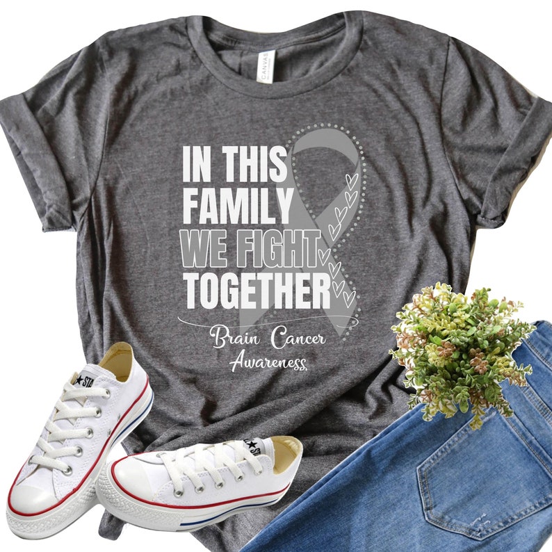 K&ouml;nnte beinhalten: Graues T-Shirt mit einem wei&szlig;en Band und dem Text "In dieser Familie k&auml;mpfen wir zusammen" und "Gehirntumor-Bewusstsein".
