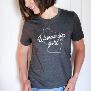 K&ouml;nnte beinhalten: Ein dunkelgraues T-Shirt mit einem wei&szlig;en Umriss des Bundesstaates Wisconsin und dem Text "Wisconsin girl" in wei&szlig;er kursivschrift.