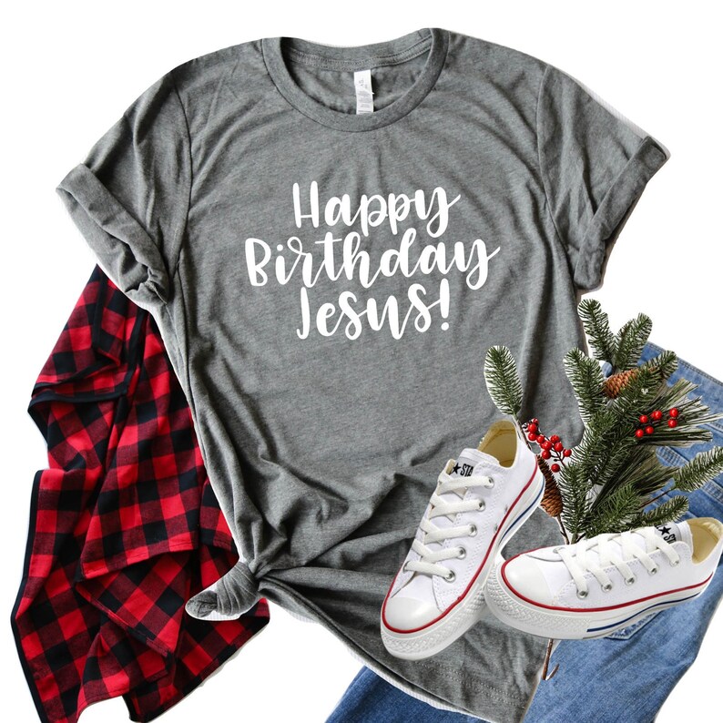 以下が含まれることがあります： 白い文字で「Happy Birthday Jesus!」と書かれたグレーのTシャツ。青いジーンズ、赤と黒のチェック柄のフランネルシャツ、白いスニーカー、赤い実のついた松の枝と合わせています。